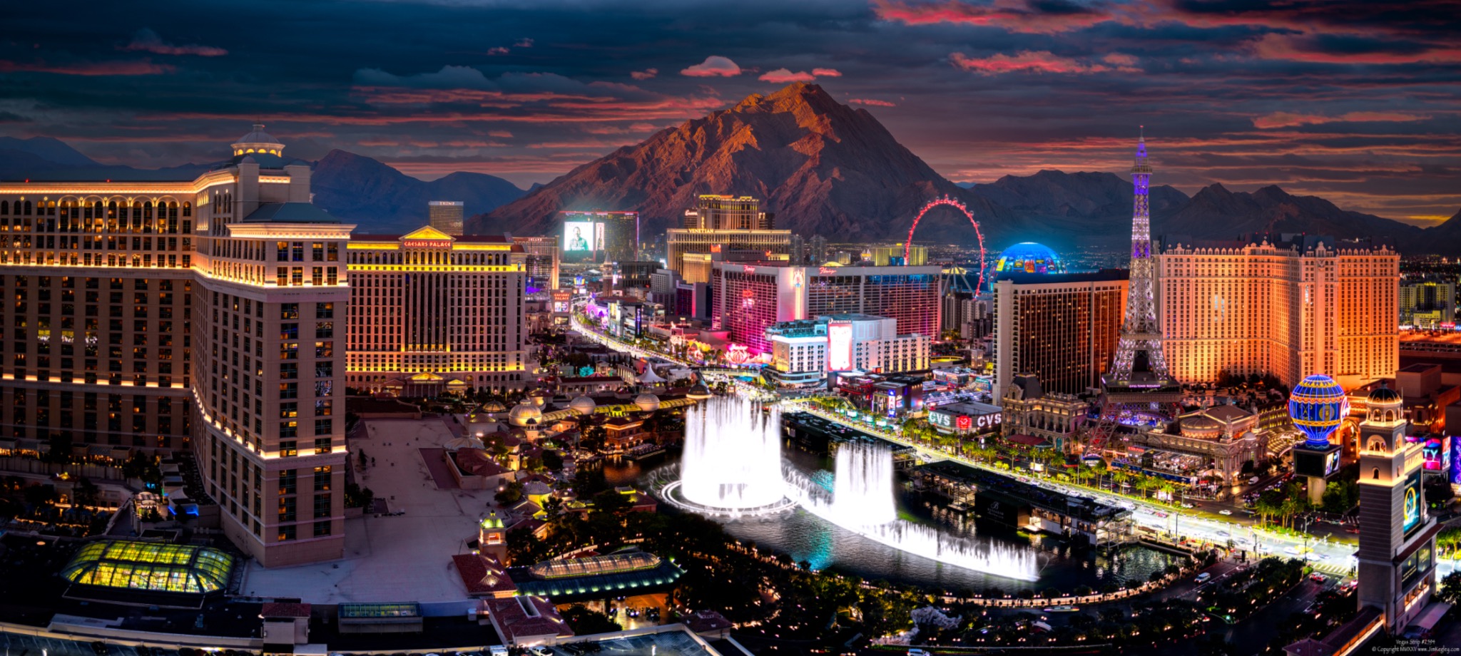 The Las Vegas Strip and Bellagio fountains at dusk, with illuminated resorts, Sphere and a mountain backdrop.